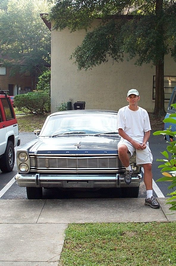 Person leaning on the front bumper of a weathered dark blue full-size Ford hardtop with stacked headlights