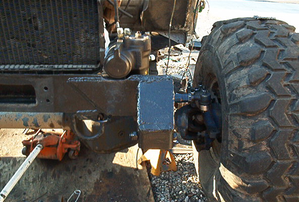 Close-up of a large off-road tire, steering linkage, and part of a radiator assembly supported by jacks