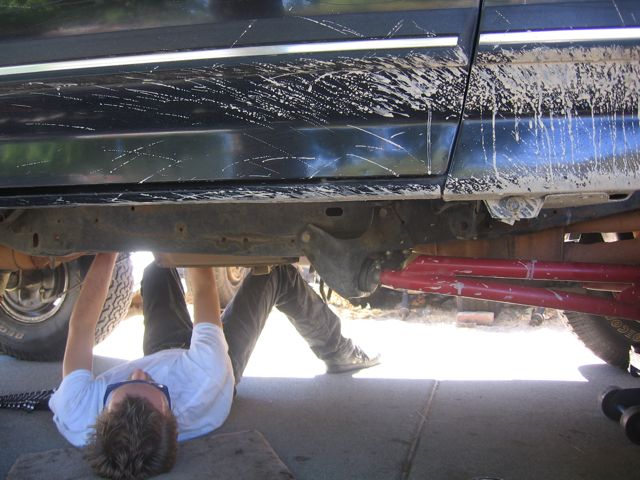 Person lying under a lifted black Ford truck working on the underbody in a driveway