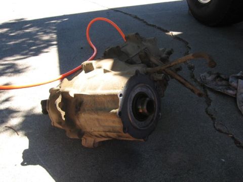 1989 Ford Bronco - A transfer case, likely removed for repair or upgrade, sits on the ground next to an orange hose and some metal rods.