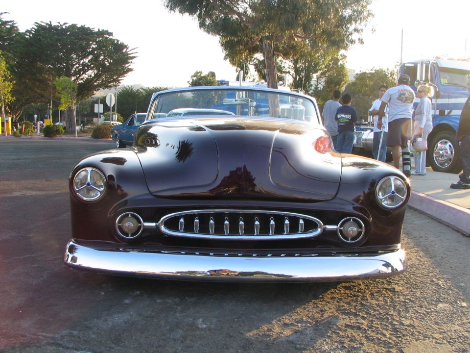 Front view of a dark purple customized hot rod with a prominent chrome grille and hood scoop. Other vehicles and people are visible in the background