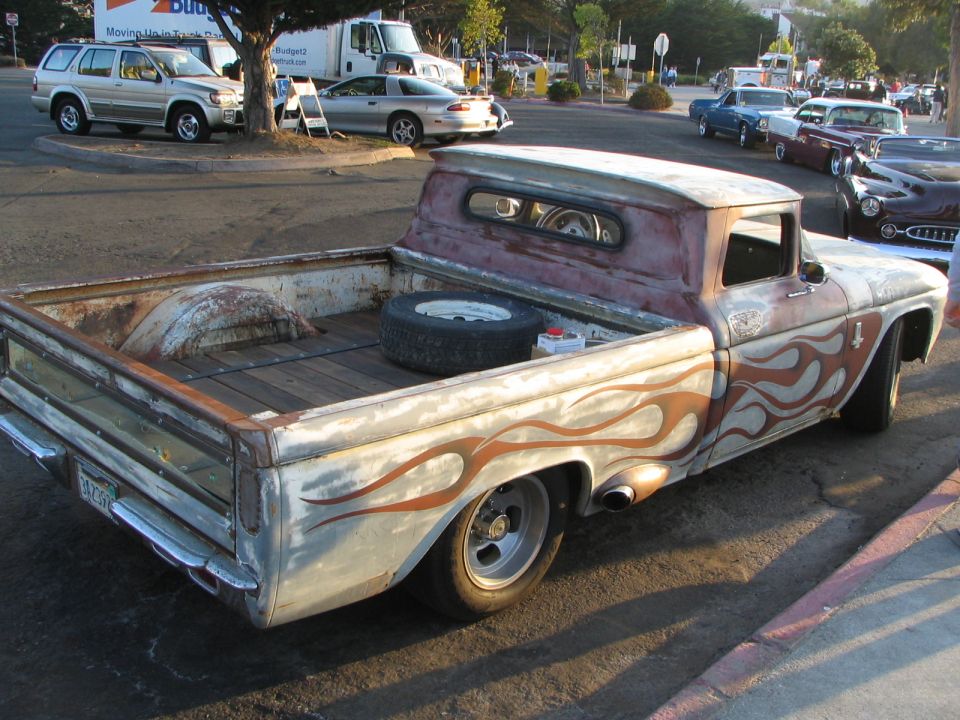 Old pickup truck with flame graphics and a spare tire in the bed. Other cars and a truck are in the background
