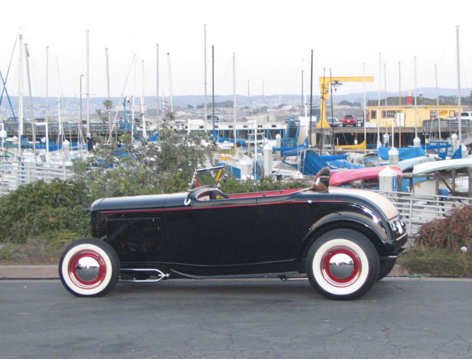 Black hot rod roadster with red trim and whitewall tires parked by a marina
