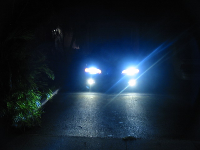 Two cars driving at night, their headlights are very bright. One car is on the left, the other on the right