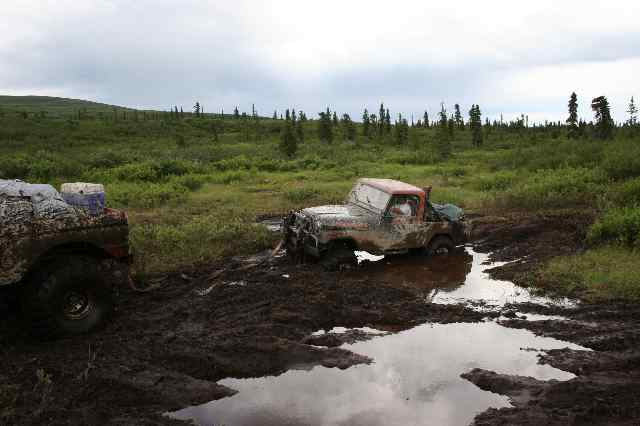 Two muddy off-road vehicles stuck in a bog with trees and green hills in the background