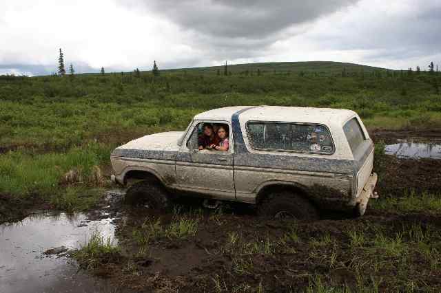 Muddy 1978 Ford Bronco with two people inside stuck in a muddy field
