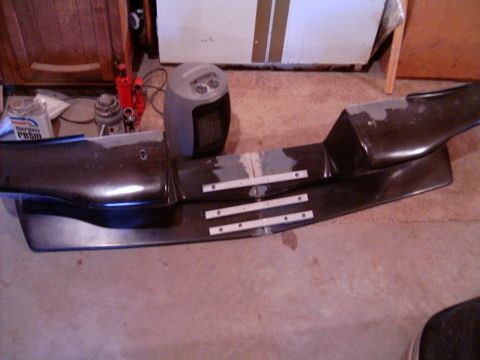 1978 Ford Mustang II - A dark fiberglass front air dam/spoiler lies face-up on a carpeted garage floor, spliced together at the center with three perforated metal…