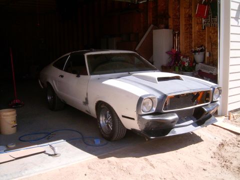 1978 Ford Mustang II - Three-quarter front view of a white Mustang II hatchback in primer sitting half out of an open garage, with a hood scoop, blacked-out front…