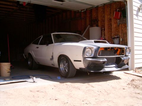 1978 Ford Mustang II - White Mustang II hatchback project car sitting in the open doorway of an unfinished stud-wall garage, shot from a low 3/4 front angle with…