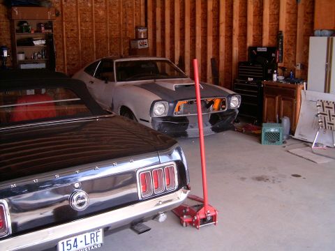 1978 Ford Mustang II - Two Mustangs share a bare-stud garage: in the foreground the black rear end of a first-generation fastback with MUSTANG lettering, tri-bar…