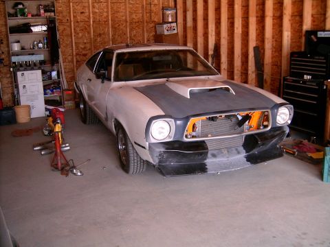 1978 Ford Mustang II - A hatchback project car sits in a wood-framed garage in primer and bare bodywork, with a hood scoop, missing grille exposing the intercoole…