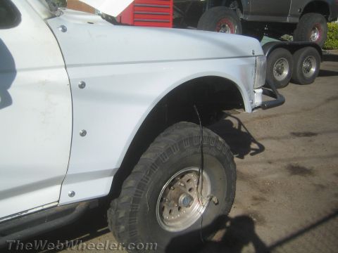 1990 Ford Bronco - Close-up of a white vehicle's front fender and off-road tire on a chrome wheel, with a truck on a trailer behind.
