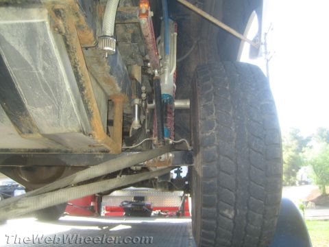 1990 Ford Bronco - Low-angle view beneath a lifted vehicle showing the rusty frame, coilover shock, steering linkage and a large tire.
