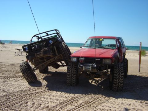 1992 Ford Bronco - Two heavily modified off-road vehicles are parked on a sandy beach with the ocean in the background.
