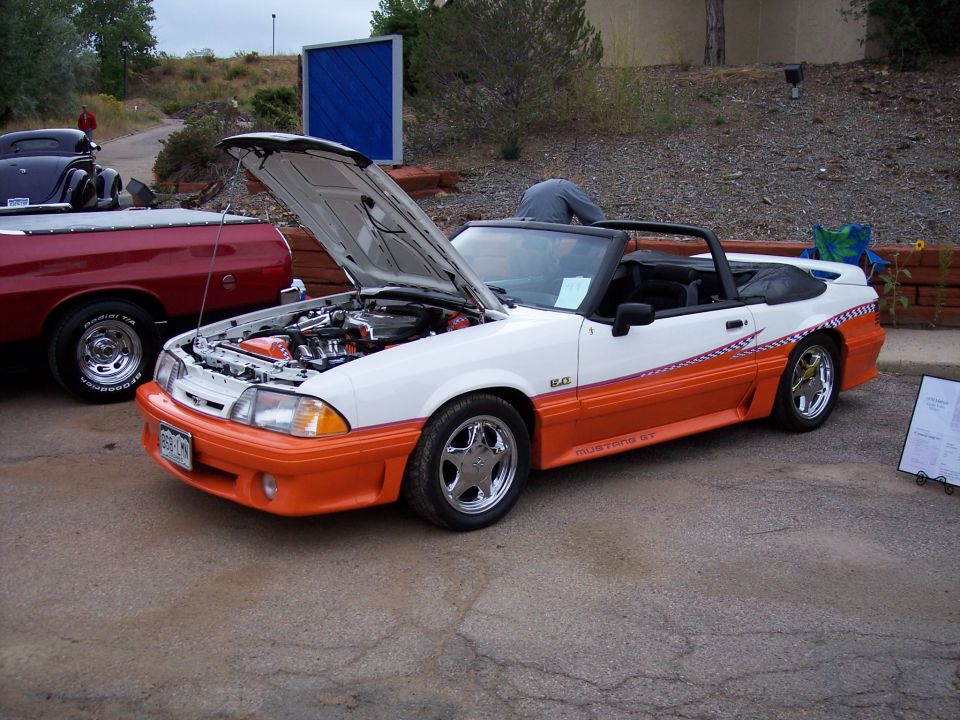 White and orange convertible Mustang GT with hood open, parked next to a red pickup truck