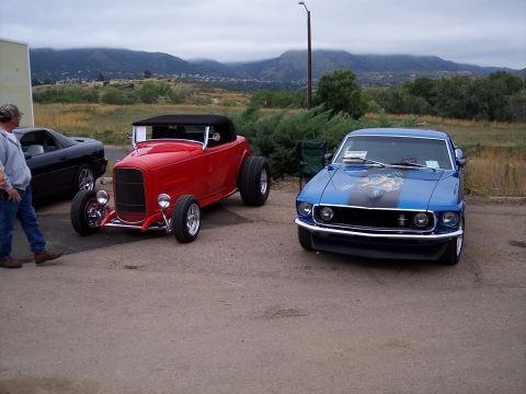1972 Ford Ranchero - Two classic cars, a blue 1969 Ford Mustang and a red hot rod, are parked outside with mountains in the background.