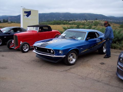 1972 Ford Ranchero - A blue 1969 Ford Mustang Mach 1 is parked next to a red hot rod and other vehicles.