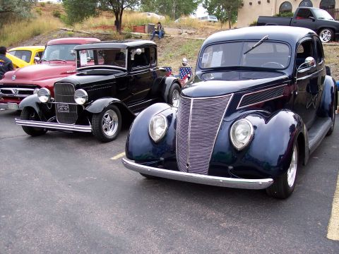 1972 Ford Ranchero - A dark blue 1937 Ford coupe is parked next to a black hot rod, with other vehicles visible in the background.