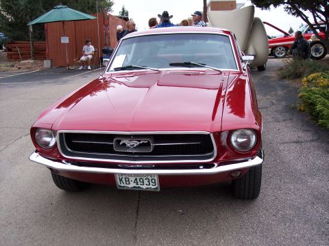 1972 Ford Ranchero - A shiny red 1967 Ford Mustang is parked at an outdoor event, with other vehicles and people in the background.