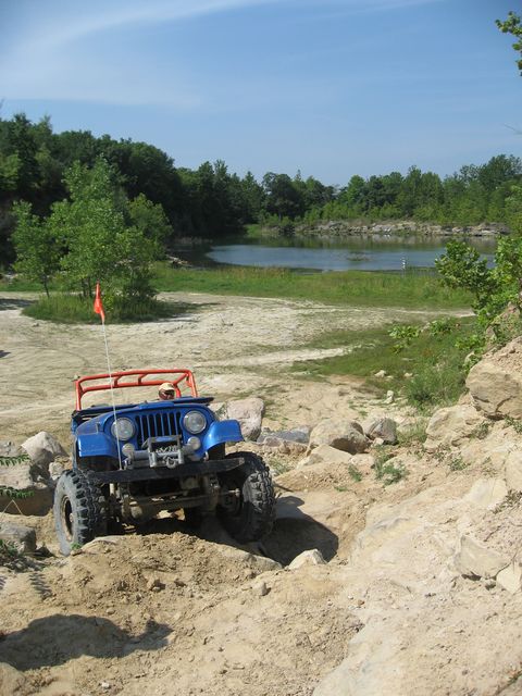 1992 Ford Bronco - A blue Jeep navigates a rocky off-road course with a body of water in the background.