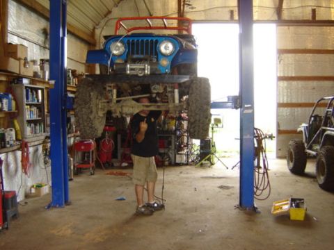 1992 Ford Bronco - A blue Jeep CJ-7 is raised on a lift in a garage, with a mechanic working on its underside. Another vehicle is parked to the side.