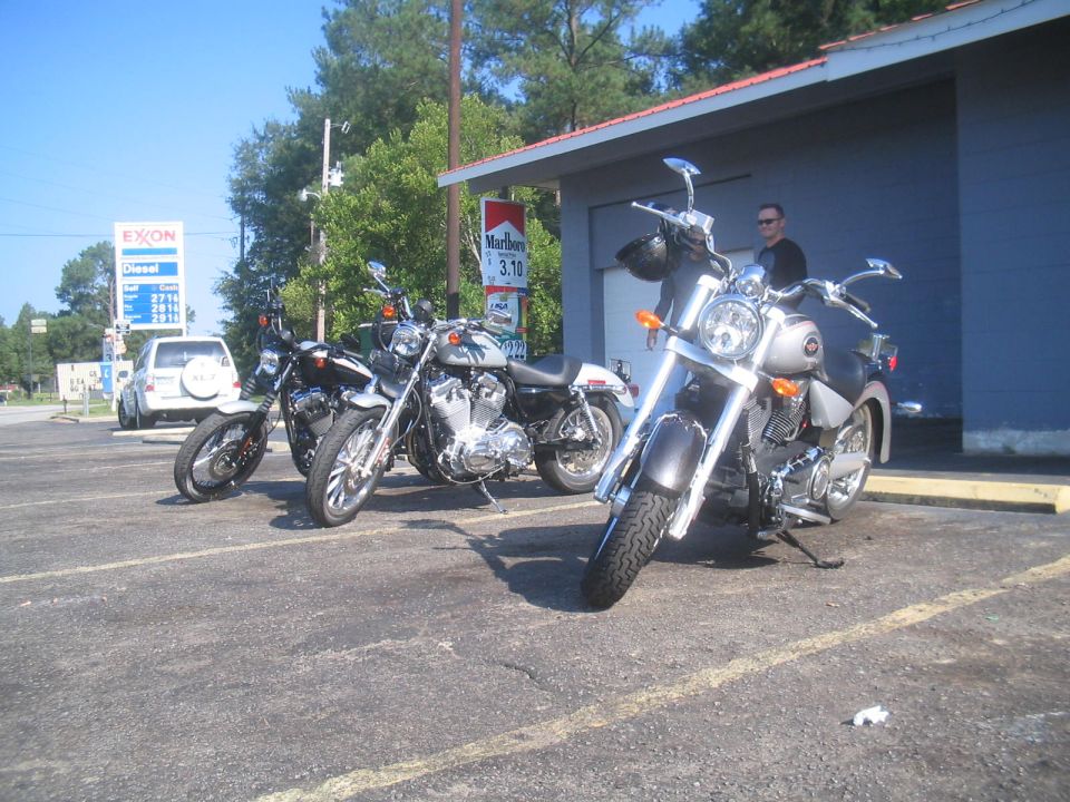 Silver motorcycle parked outside a building with other motorcycles and a gas station in the background