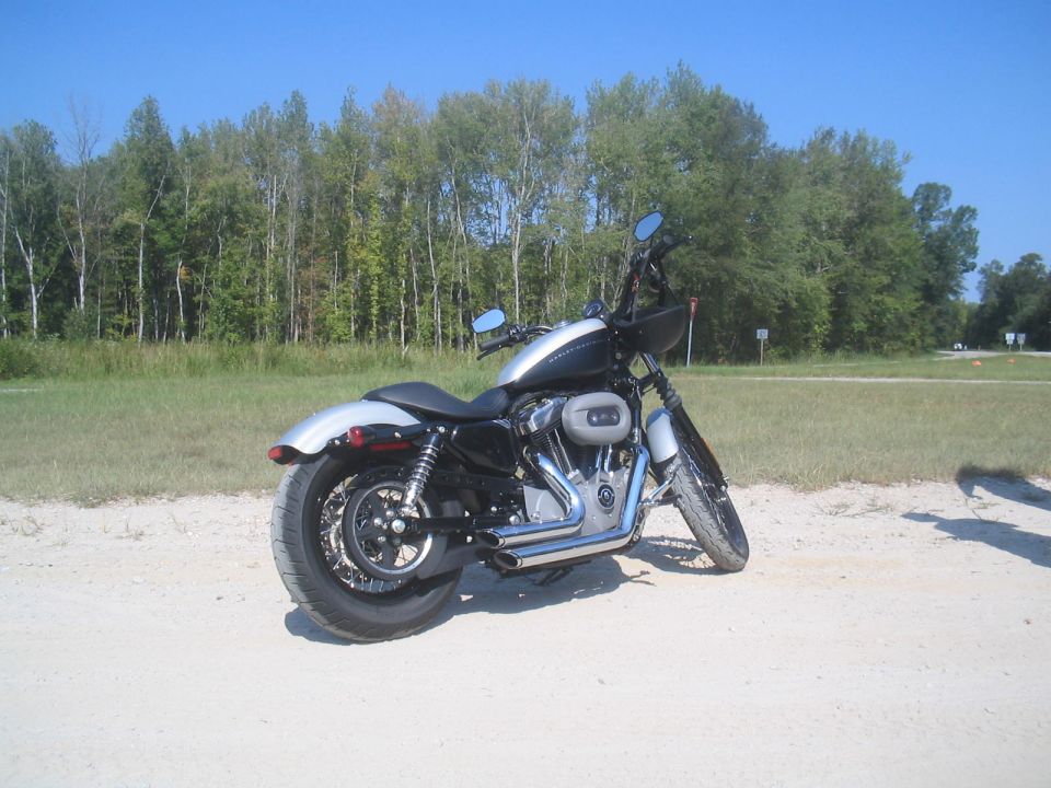 Harley-Davidson motorcycle parked on dirt road with trees in background. Watermark says SUPERMOTORS.NET
