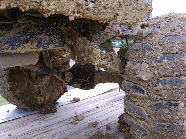 Mud-caked solid front axle and tires under a lifted off-road truck on a wooden ramp