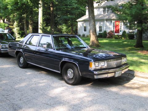 1991 Ford LTD Crown Victoria - dark blue four-door Ford sedan parked front-quarter on a driveway before a white house with a banner.