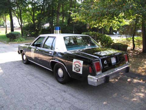 1991 Ford LTD Crown Victoria - Rear three-quarter of a black-and-white police sedan marked "POLICE" and an Emergency 911 door decal, on a driveway.