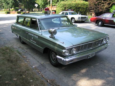 1991 Ford LTD Crown Victoria - green "Stratford Police Radio Patrol" station wagon with roof beacon and hood siren parked on a driveway near other cars.