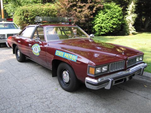 1991 Ford LTD Crown Victoria - maroon four-door sedan lettered STATE PATROL with a star door emblem and roof lights is parked on asphalt near greenery.