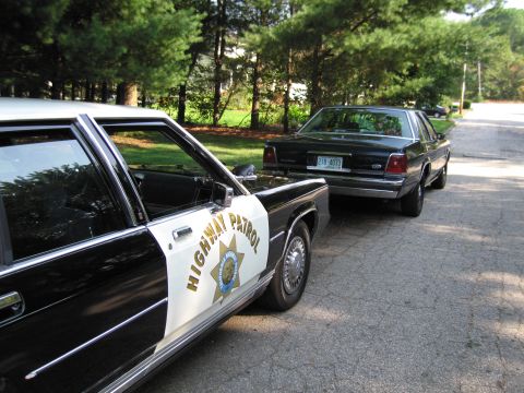 1991 Ford LTD Crown Victoria - black-and-white HIGHWAY PATROL cruiser with a gold star door shield sits beside a dark sedan on a quiet street.