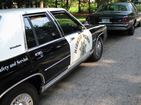 1991 Ford LTD Crown Victoria - Close view of a black-and-white HIGHWAY PATROL cruiser door with a star shield and the words Safety and Service, next to a dark Ford sedan.