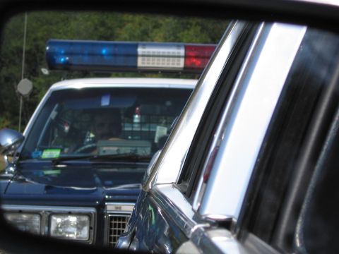 1991 Ford LTD Crown Victoria - police car's roof lightbar and front grille are framed through the window of another vehicle, with a driver visible at the wheel.