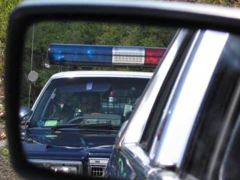 1991 Ford LTD Crown Victoria - police cruiser with a red-and-blue roof lightbar and a driver at the wheel is captured in a vehicle's side mirror.