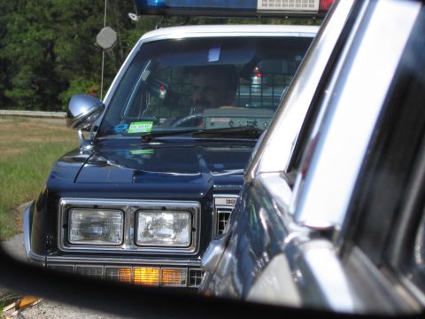 1991 Ford LTD Crown Victoria - dark police sedan with rectangular headlights and a roof lightbar follows closely, its driver visible through the windshield.