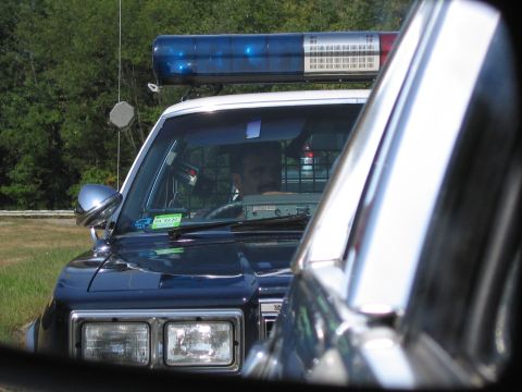 1991 Ford LTD Crown Victoria - dark police car with a blue roof lightbar and a driver at the wheel is framed through the windshield of the car ahead.