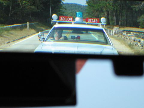 1991 Ford LTD Crown Victoria - white STATE POLICE car with red roof light boxes approaches head-on on a country road, seen through a rear window.