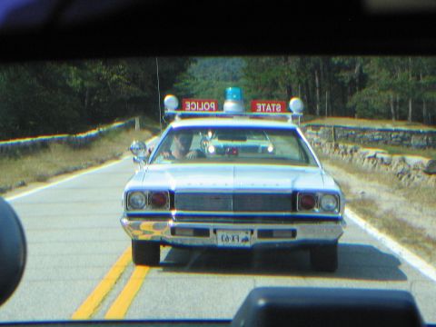 1991 Ford LTD Crown Victoria - white police car displaying STATE POLICE roof light boxes drives toward the camera on a stone-walled two-lane road.