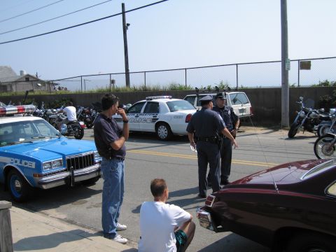 1991 Ford LTD Crown Victoria - Men in casual clothes and uniformed officers gather beside a blue POLICE sedan and a white cruiser at a fenced beachside lot.