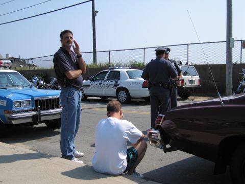 1991 Ford LTD Crown Victoria - man stands and another sits on the curb beside a blue police car and a white cruiser as an officer looks on in a fenced lot.
