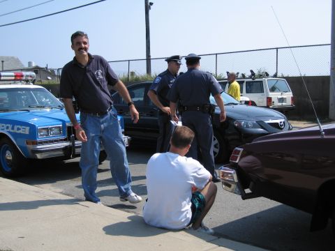 1991 Ford LTD Crown Victoria - smiling mustached man in a dark polo stands beside a blue POLICE sedan while officers talk near other vehicles in a fenced lot.