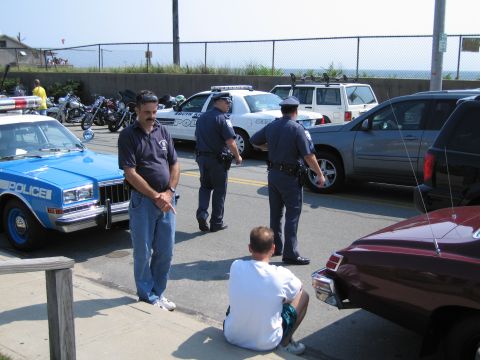 1991 Ford LTD Crown Victoria - man in a dark polo stands by a blue POLICE sedan as two officers converse near a white cruiser and SUV in a fenced beachside lot.
