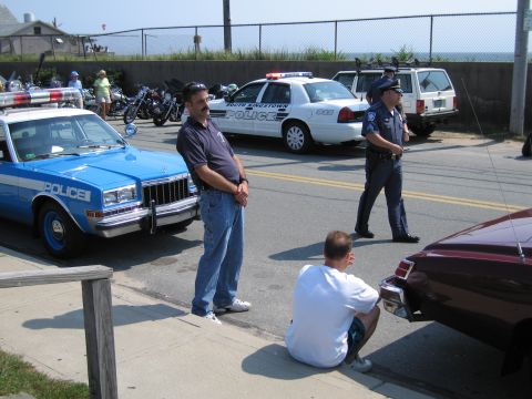 1991 Ford LTD Crown Victoria - man stands beside a blue POLICE sedan while a uniformed officer and a white police cruiser occupy a fenced beachside parking lot.