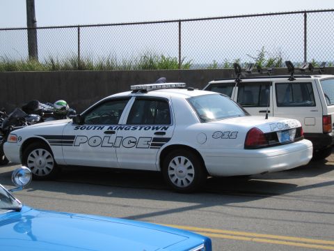 1991 Ford LTD Crown Victoria - white SOUTH KINGSTOWN POLICE cruiser marked 911 with a roof lightbar is parked beside an SUV at a fenced beachside lot.