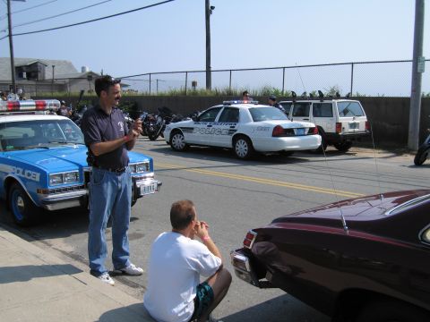 1991 Ford LTD Crown Victoria - man takes a photo while another stands by a blue POLICE sedan, with a white SOUTH KINGSTOWN cruiser and SUV across the lot.