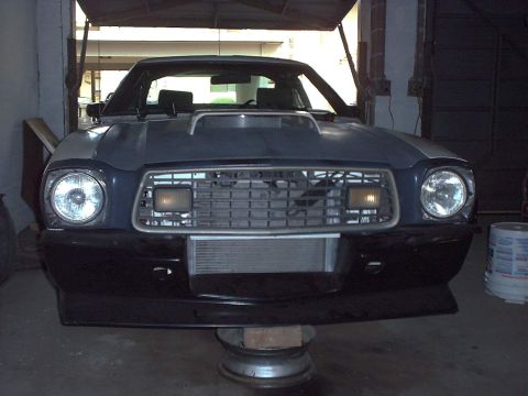 1978 Ford Mustang II - Head-on front view of a dark blue/black Mustang II hatchback sitting in an open garage bay, with an egg-crate grille, round quad-style head…