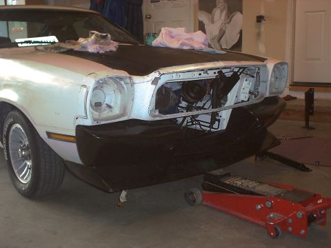 1978 Ford Mustang II - Front three-quarter view of a white Mustang II with a black hood, torn down in a home garage: the grille and headlight bezels are removed,…