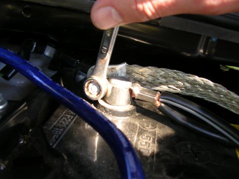 2007 Mazda 3 - A close-up shows a wrench being used to secure a car battery terminal, connecting a braided ground strap and electrical wires.